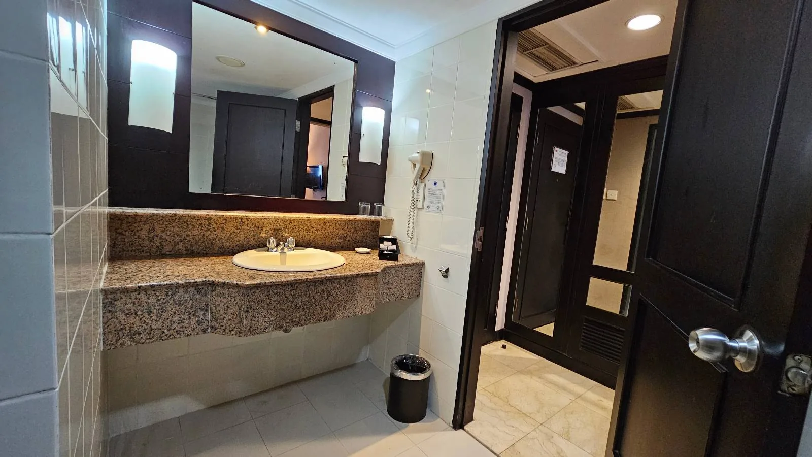 The image shows a modern bathroom with a granite countertop, a built-in sink, and a mirror with lights on either side. A hairdryer is mounted on the wall, and there's an open door leading to another room.