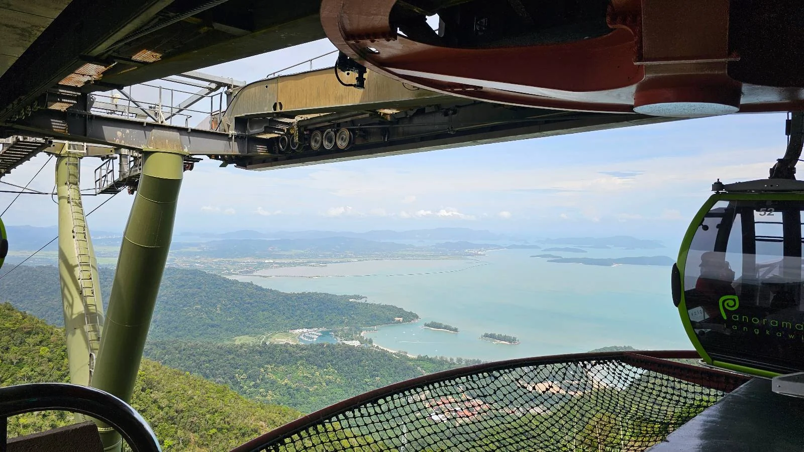 A scenic view from a cable car station overlooking a vast expanse of turquoise sea and distant land. The station structure and a cable car pod are visible in the foreground, with lush green hills below. The sky is clear with scattered clouds.