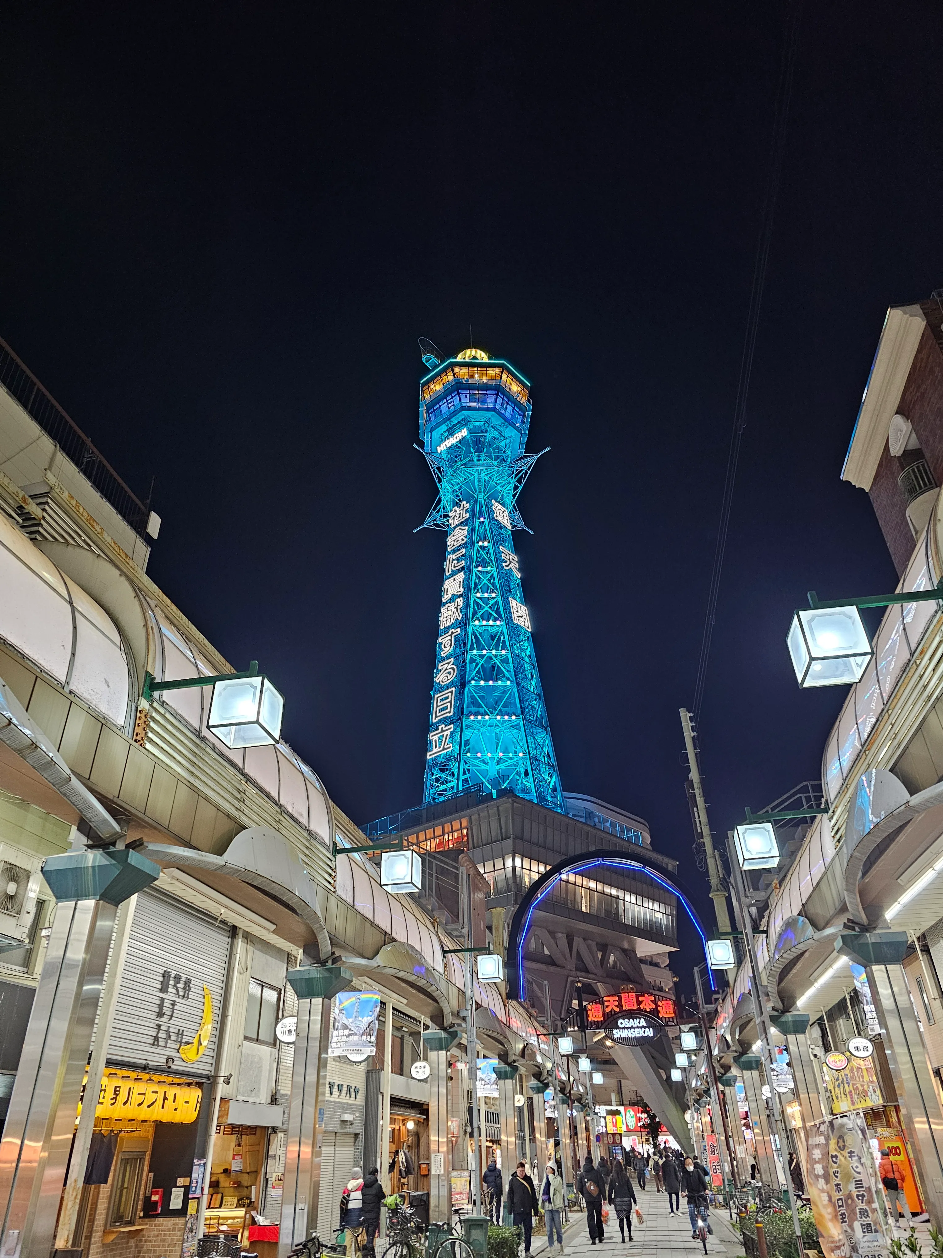 A brightly lit street scene at night in Osaka, Japan, with the tall, illuminated Tsutenkaku Tower glowing blue in the background and people walking along the lively shopping street below.