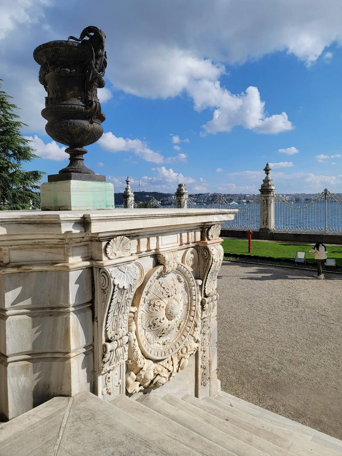Ornate stone railing with carved details and a large urn overlooks a waterfront and cityscape under a blue sky with scattered clouds. Green grass and stone posts are visible in the background.