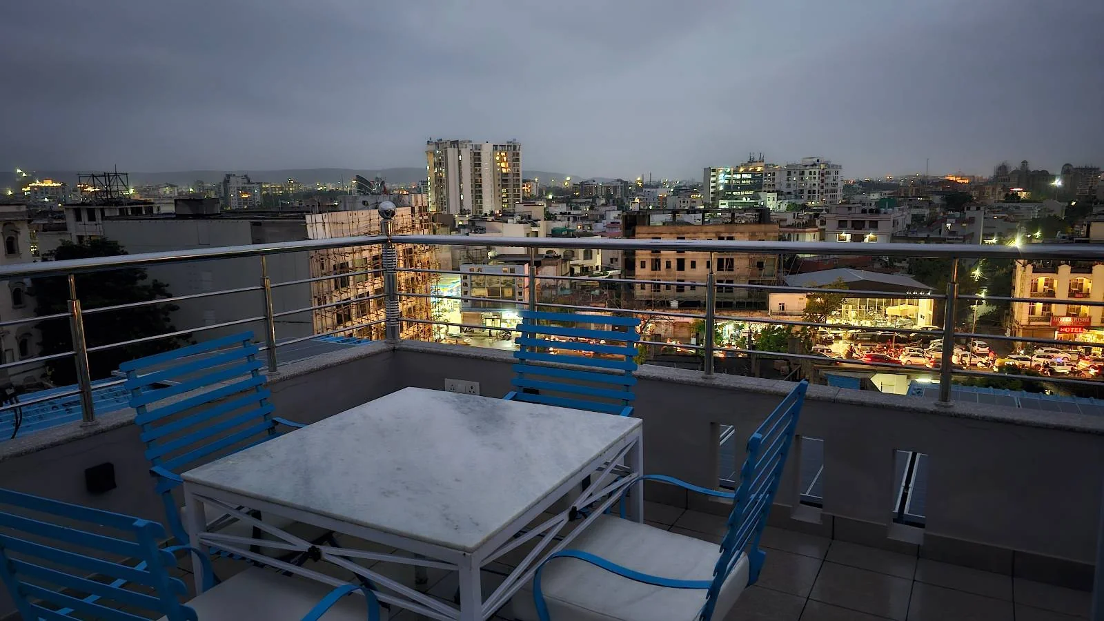 A rooftop patio with a white table and blue chairs overlooks a cityscape at dusk. Tall buildings and city lights are visible in the background under a cloudy evening sky.