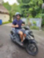 A person with sunglasses and a helmet gives a thumbs-up while sitting on a black scooter. Lush greenery and a "HIATUS" sign are in the background.