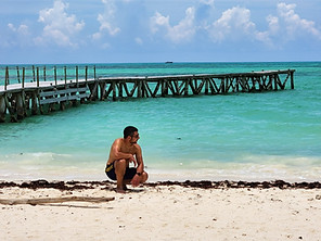 A person squats on a sandy beach near the shore in Mexico, with turquoise water and a wooden pier extending into the sea under a partly cloudy sky. Seaweed is scattered along the sand, painting a picture of stunning beaches often found near ancient ruins.
