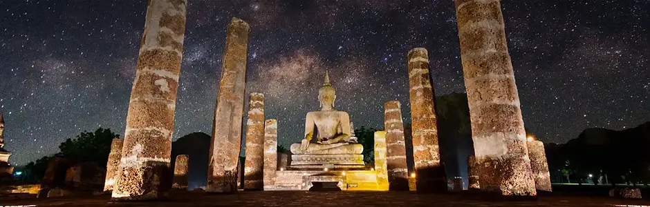 A wide panoramic shot at night features a large, ancient stone Buddha statue seated in a meditative pose. The statue sits centered on a raised platform framed by weathered stone pillars. A brilliant, star-filled night sky with a visible Milky Way galaxy glows intensely in the background.
