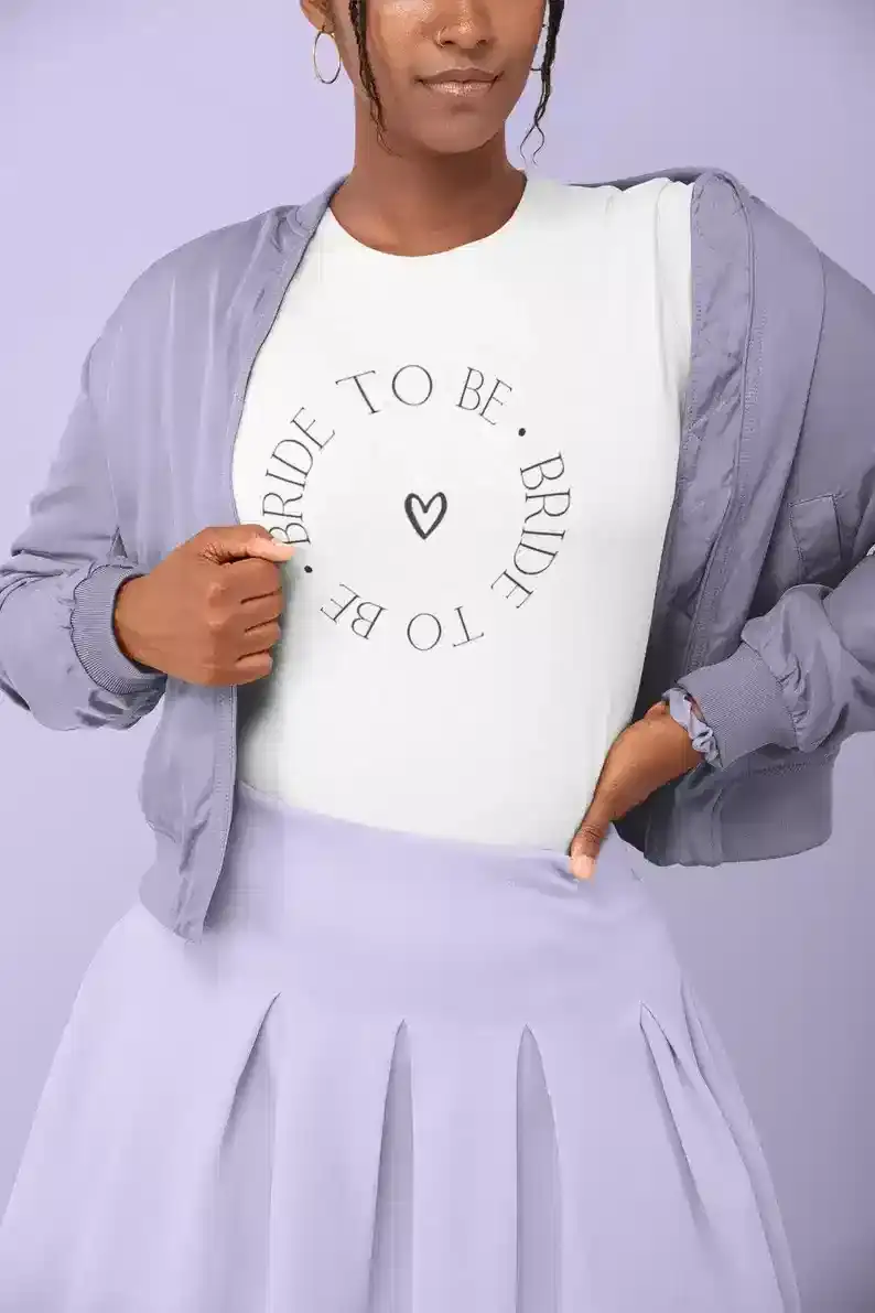 A person wearing a white shirt with "BRIDE TO BE" printed in a circle around a small heart, paired with a light purple jacket