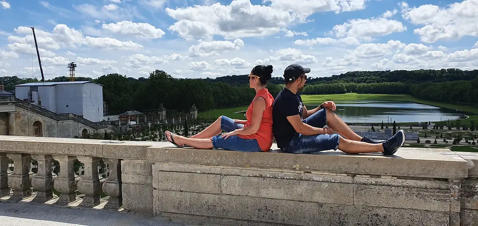 Two people in casual clothes sit back-to-back on a stone ledge overlooking a scenic park and lake under a blue sky with clouds.
