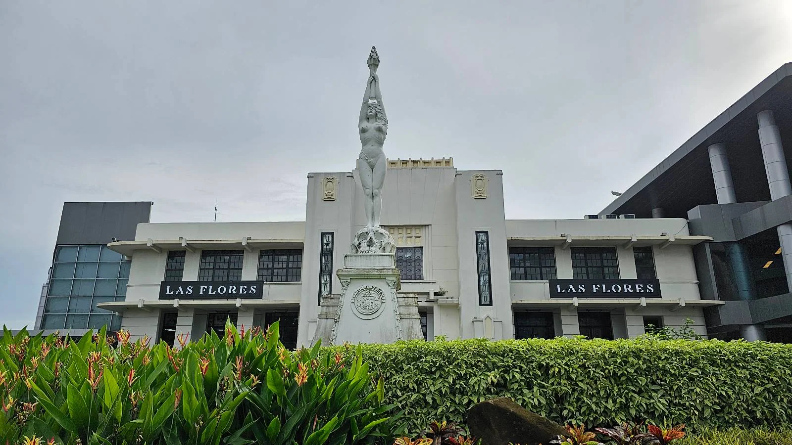 A tall white statue stands in front of a historic building with two visible signs reading "Las Flores." The building has a gray and white facade, surrounded by lush green vegetation under a cloudy sky.