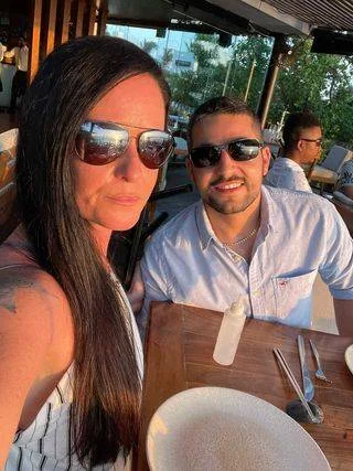 A woman and a man are sitting at an outdoor restaurant table during sunset. Both are wearing sunglasses and smiling. The table is set with plates and utensils. Trees and another person are visible in the background.