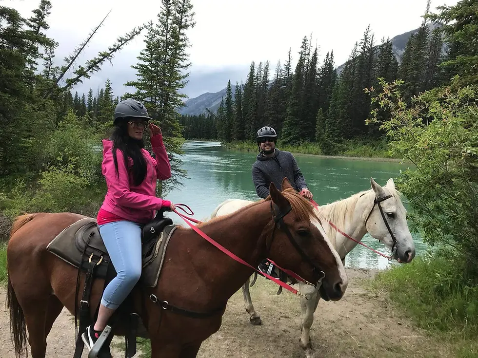 Two people riding horses near a turquoise lake, surrounded by evergreen trees and mountains.