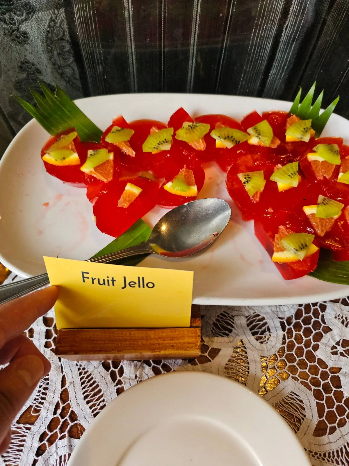 A white platter with red fruit jelly pieces topped with yellow fruit cubes, garnished with green leaves. A small sign in front reads "Fruit Jelly." The platter sits on a lace tablecloth.