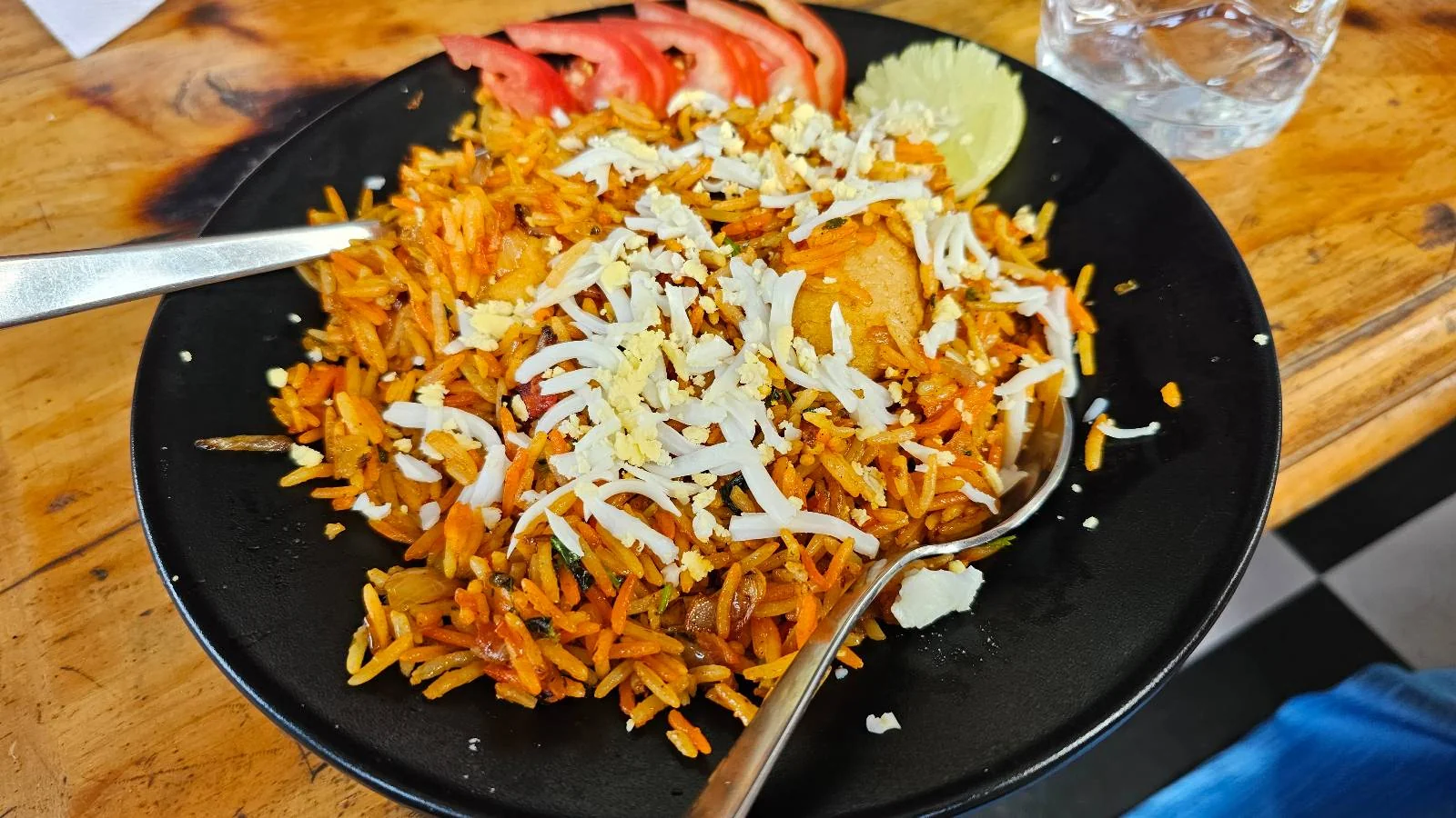 A black plate of biryani garnished with grated cheese, served with sliced tomatoes and a wedge of lime, with a fork and spoon on the side. A glass of water is visible in the background on a wooden table.
