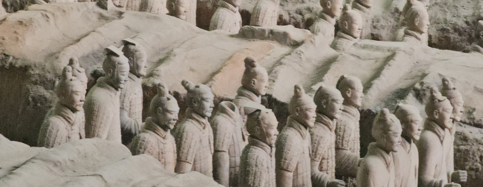 Rows of the Terracotta Army statues, partially buried in earth, stand in an archaeological excavation site in China. The life-sized soldier figures have distinct facial features and are positioned in formation.