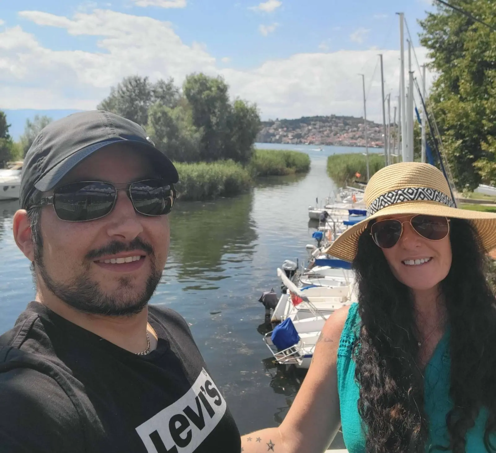 A man and woman smile, taking a selfie by a small canal lined with boats. He's wearing sunglasses and a black T-shirt. She's in a teal top with a wide-brimmed hat. In the background, trees and a distant town are visible under a partly cloudy sky.