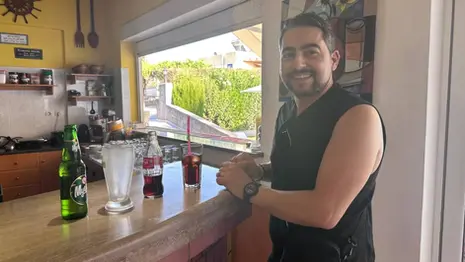 Leon standing at the bar at Michalis Apartments in Greece, with drinks on the counter and a casual indoor bar setting visible behind him.