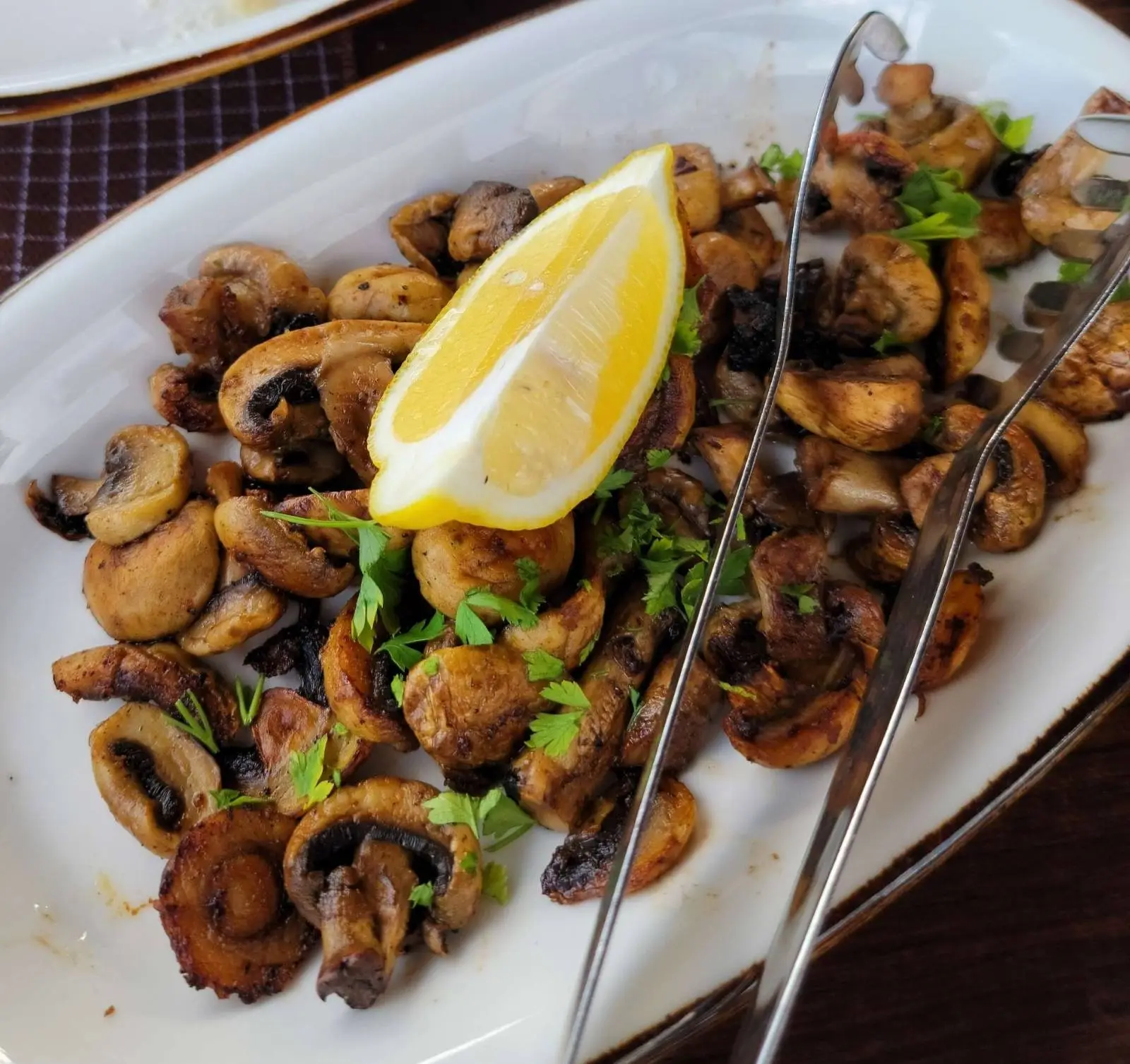 A white dish filled with sautéed mushrooms, garnished with chopped parsley and a lemon wedge on top. Silver serving tongs rest on the dish, all placed on a dark checkered tablecloth.