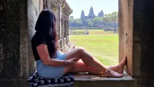 Woman in a black shirt and denim shorts sits in a stone window at Angkor Wat, gazing at the temple. Sunlit green landscape outside.