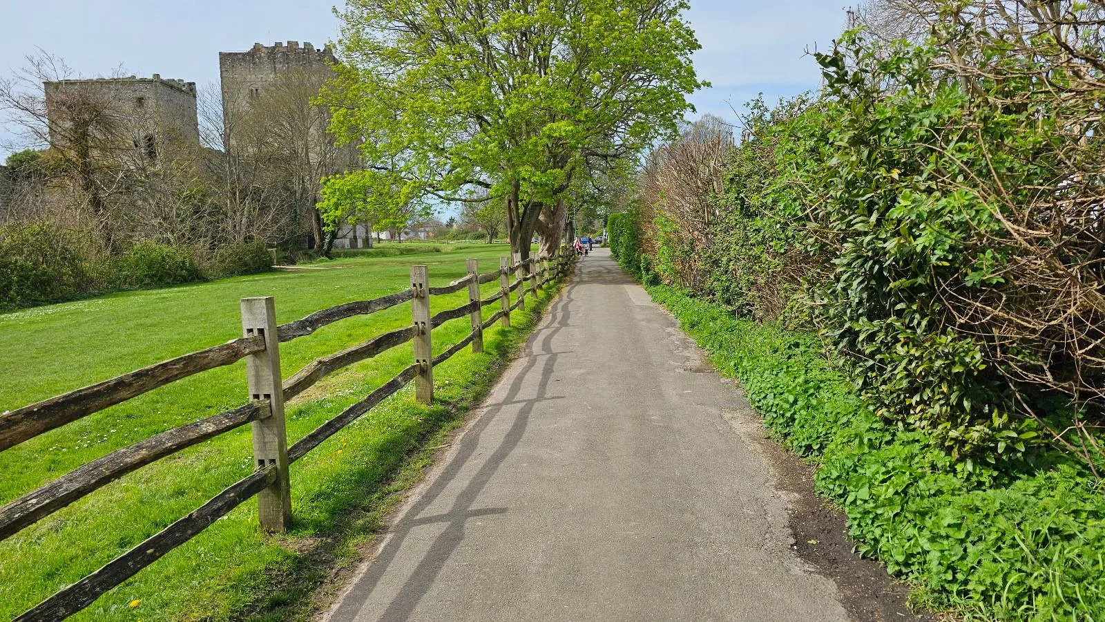 A narrow paved path lined with wooden fences and lush greenery, leading towards an ancient stone castle under a clear blue sky.