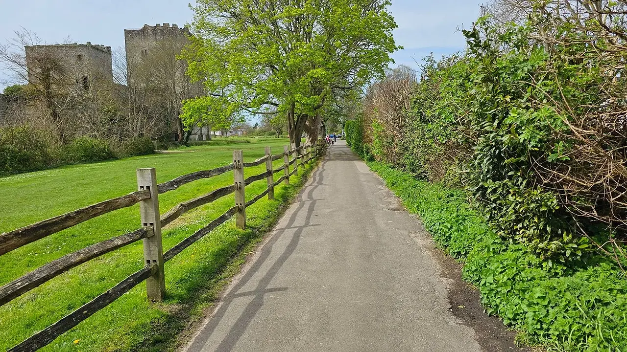A narrow paved path lined with wooden fences and lush greenery, leading towards an ancient stone castle under a clear blue sky.