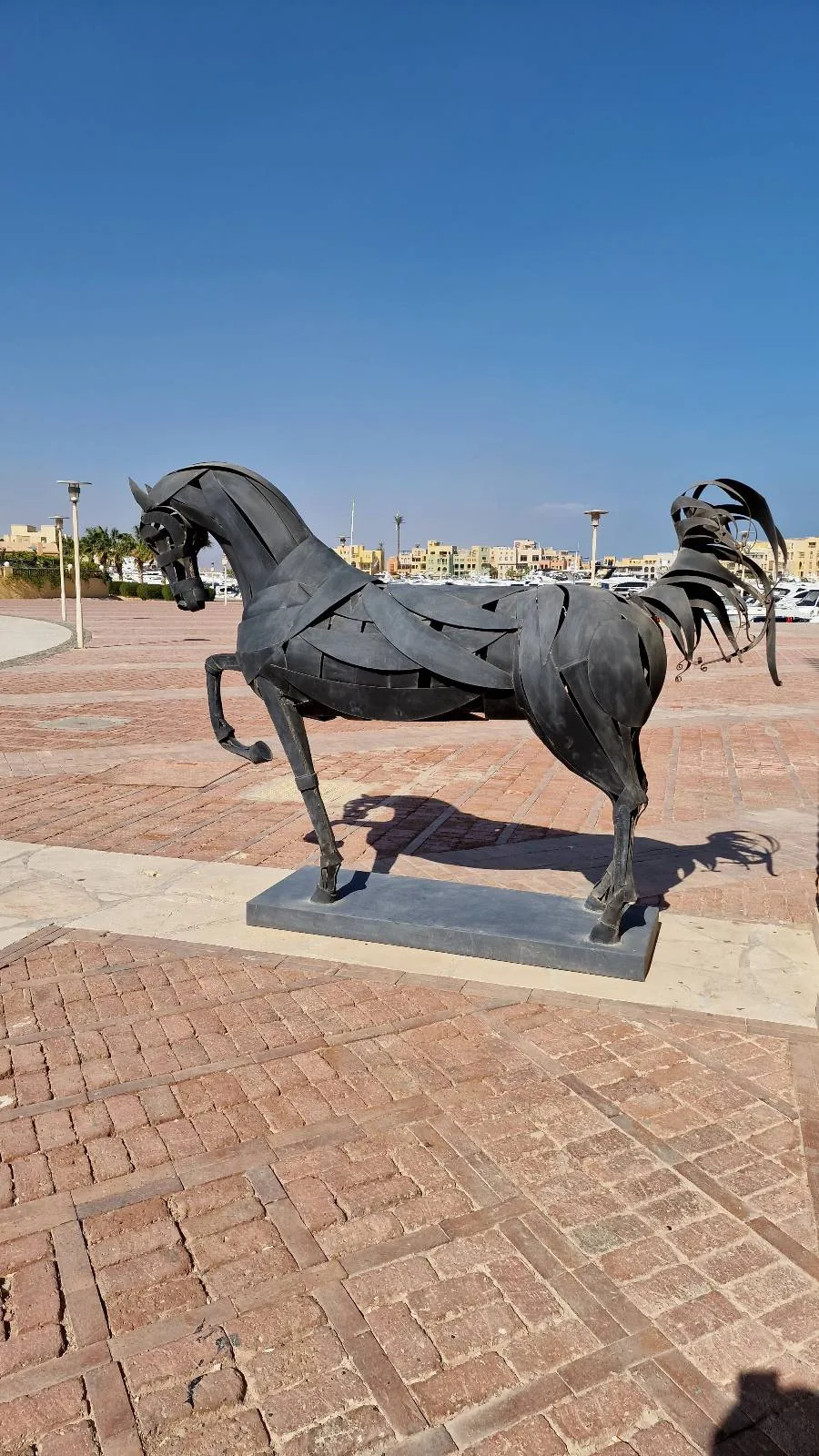 Bronze statue of a rearing horse standing on a paved area with lamp posts and buildings in the background on a clear day.