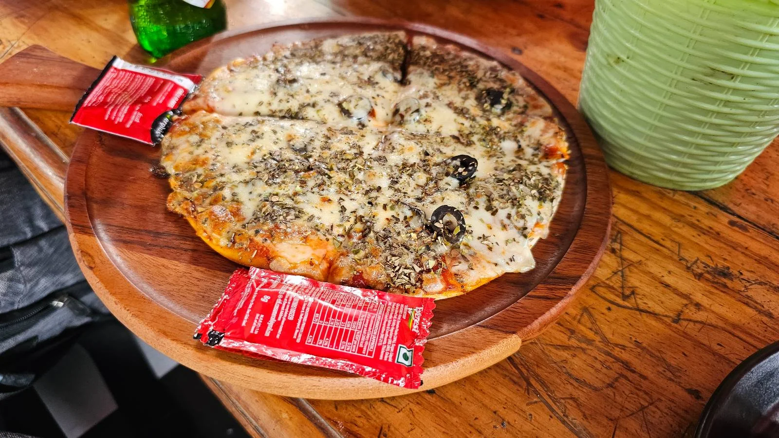 A round pizza topped with cheese, herbs, and black olives is served on a wooden platter with two red sauce packets on the side, set on a rustic wooden table next to a green container.