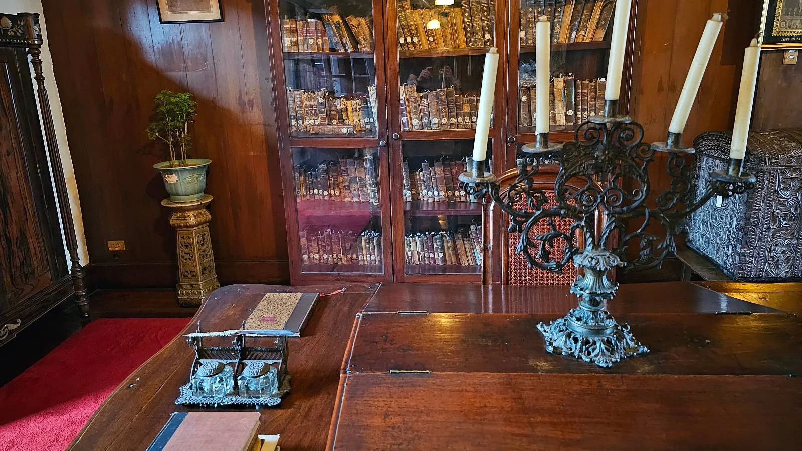 A cozy study room with a wooden desk featuring an ornate candelabra and a decorative item. A bookshelf filled with books is seen in the background, alongside a small potted plant and a wooden cabinet. The room has a warm, classic ambiance.