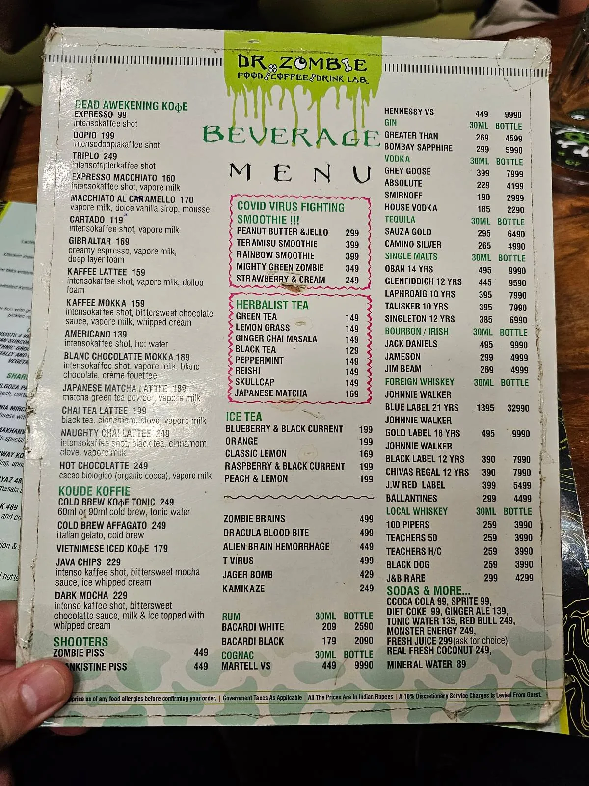 A hand holds a breakfast menu featuring a variety of food and drink options, including eggs, omelettes, pancakes, sandwiches, sides, juices, and hot beverages, with prices listed beside each item.