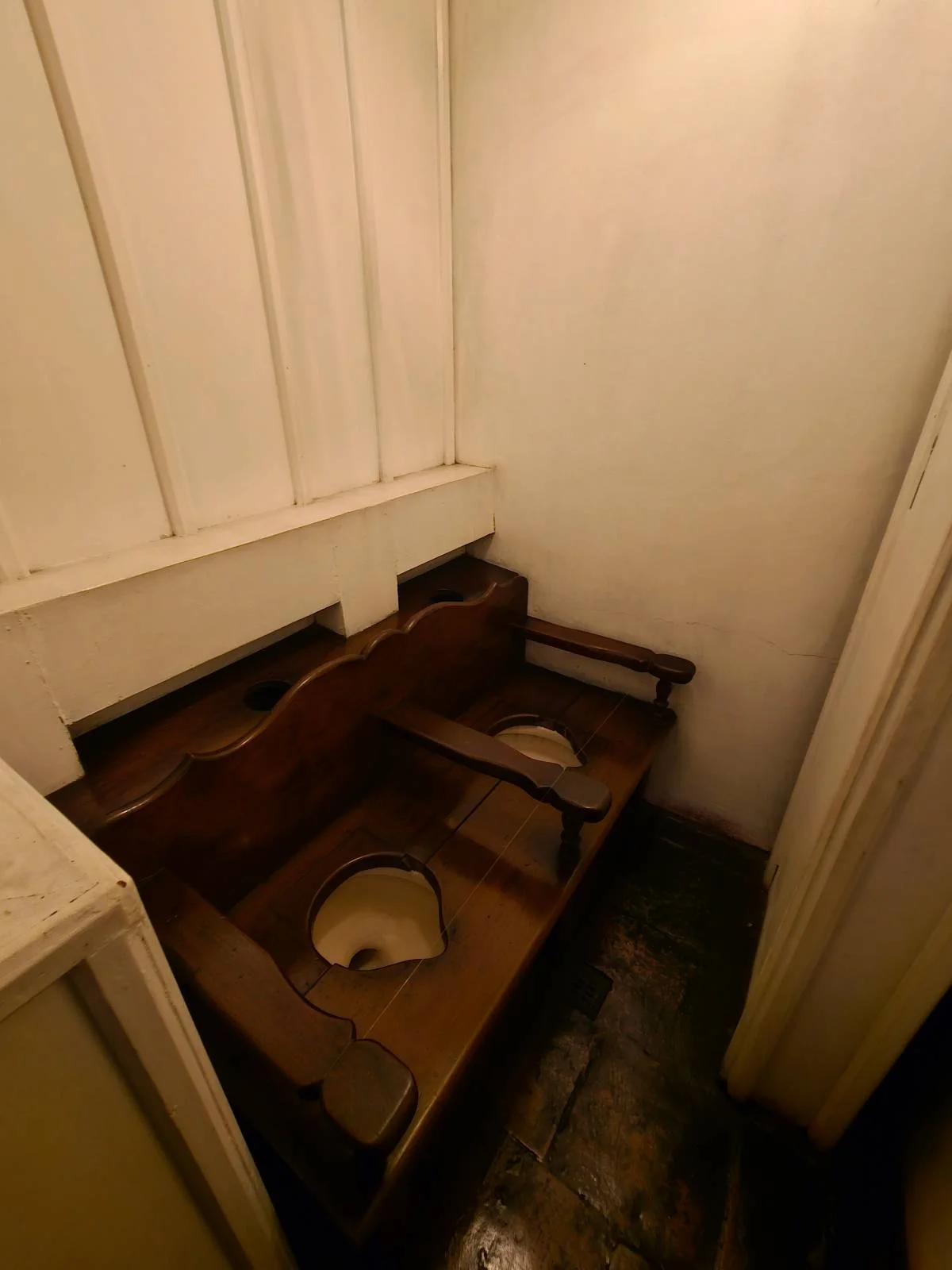 A dimly lit, rustic bathroom with two wooden toilet seats attached to a bench-like structure. The walls are white, and the floor is made of dark stone. The setting appears old-fashioned and utilitarian.