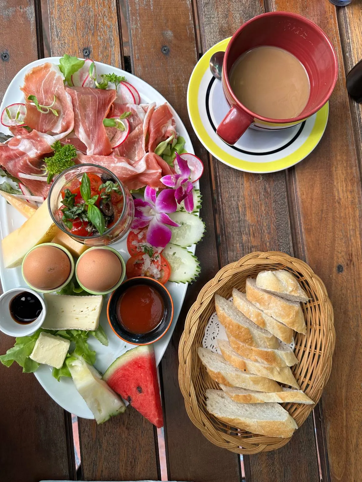 A breakfast spread on a wooden table featuring a platter with prosciutto, cheese, lettuce, sliced cucumbers, and cherry tomatoes, two soft-boiled eggs, and a basket of sliced bread. A cup of coffee rests on a saucer nearby.