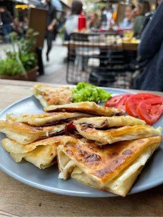 A platter with cheese, salami, cucumbers, olives, tomatoes, and jam. A bowl of bread and a drink are in the background.