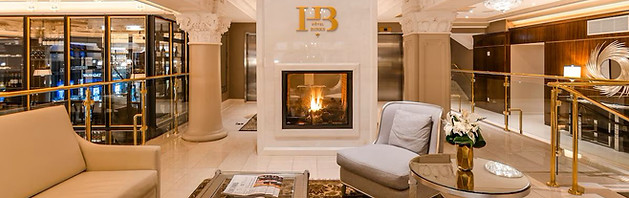 Elegant hotel lobby with a lit fireplace set in a marble wall, surrounded by plush seating. Gold railings and decorative columns accent the space, reminiscent of luxury resorts. A glass cabinet and mirror reflect the warm ambiance, while a small table holds a flower arrangement.