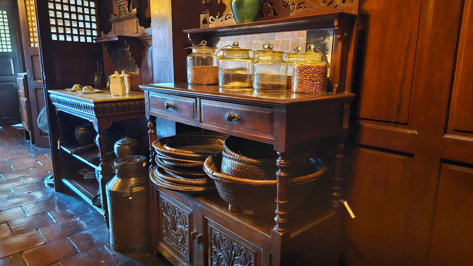 A rustic wooden kitchen cabinet holds jars filled with various ingredients. Below, large bowls and pots are neatly stacked. The warm-toned kitchen has tiled floors and wooden walls, creating a cozy, vintage atmosphere.