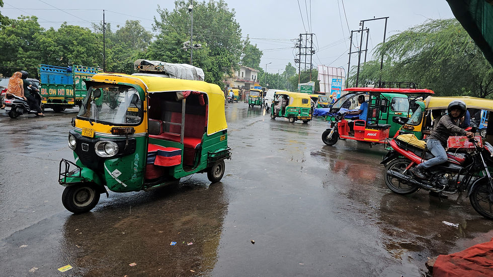 Rain-soaked street with colorful auto rickshaws and a motorcyclist. Pedestrians and blurred buildings in the background. Text reads "BLOW HORN."