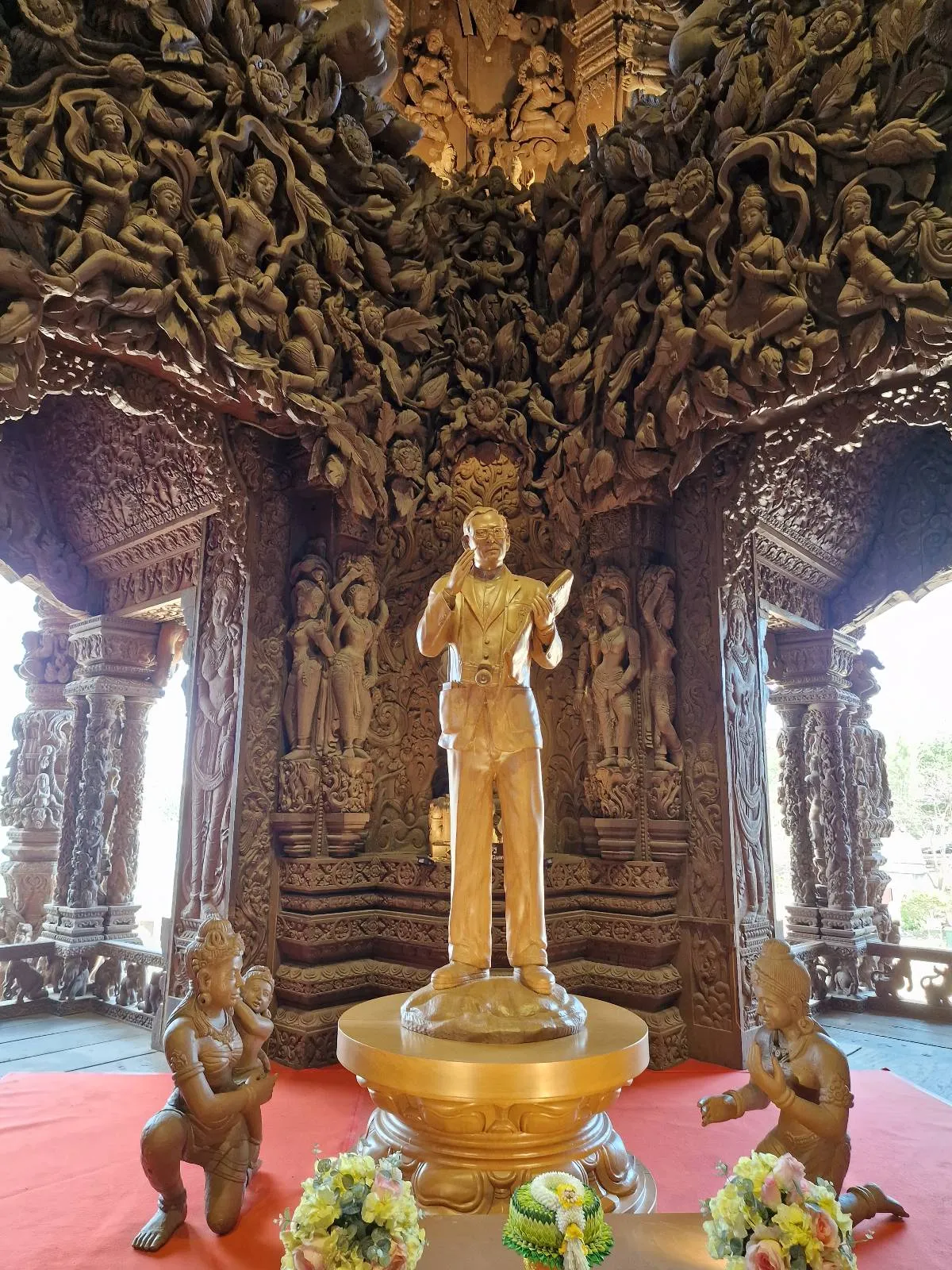 A golden statue stands on a pedestal in a richly carved wooden hall. Flanked by two smaller statues, the intricate detailing of the walls and ceiling adds depth and texture. Floral arrangements are placed in front of the display on a red carpet.