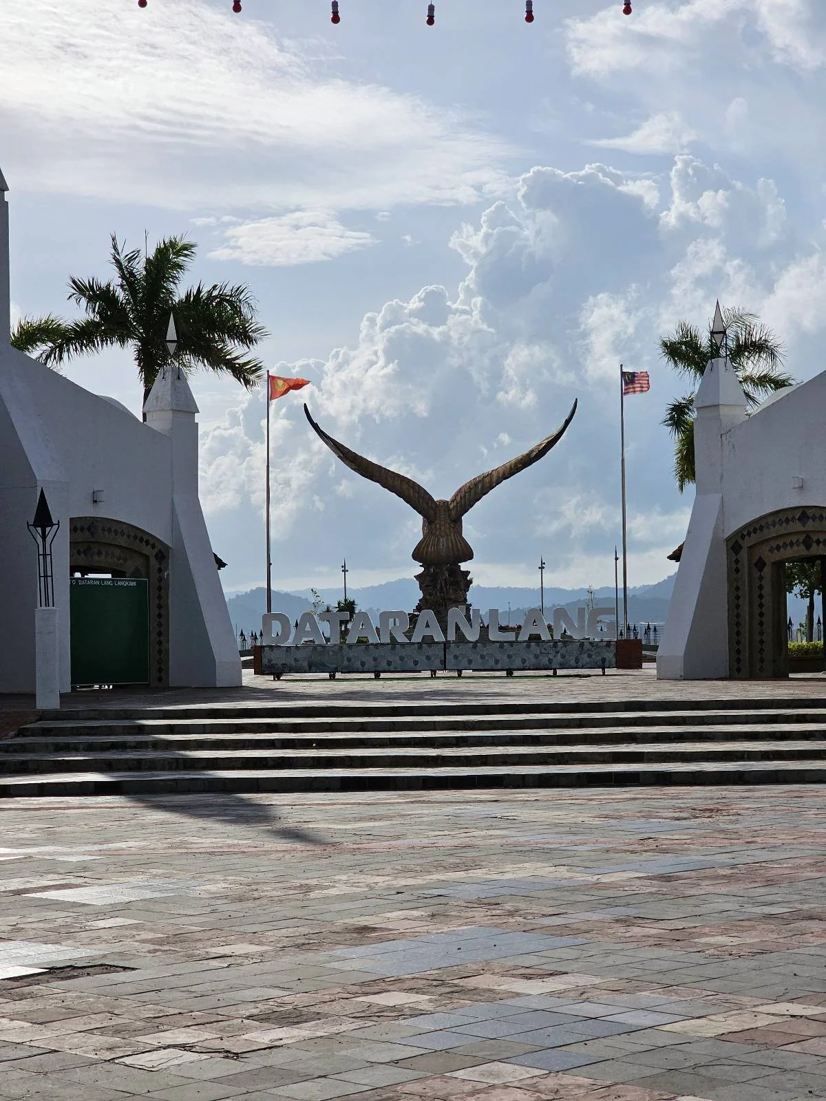 A large eagle sculpture with outstretched wings stands at a waterfront promenade, flanked by two white archways, palm trees, and flagpoles under a partly cloudy sky.