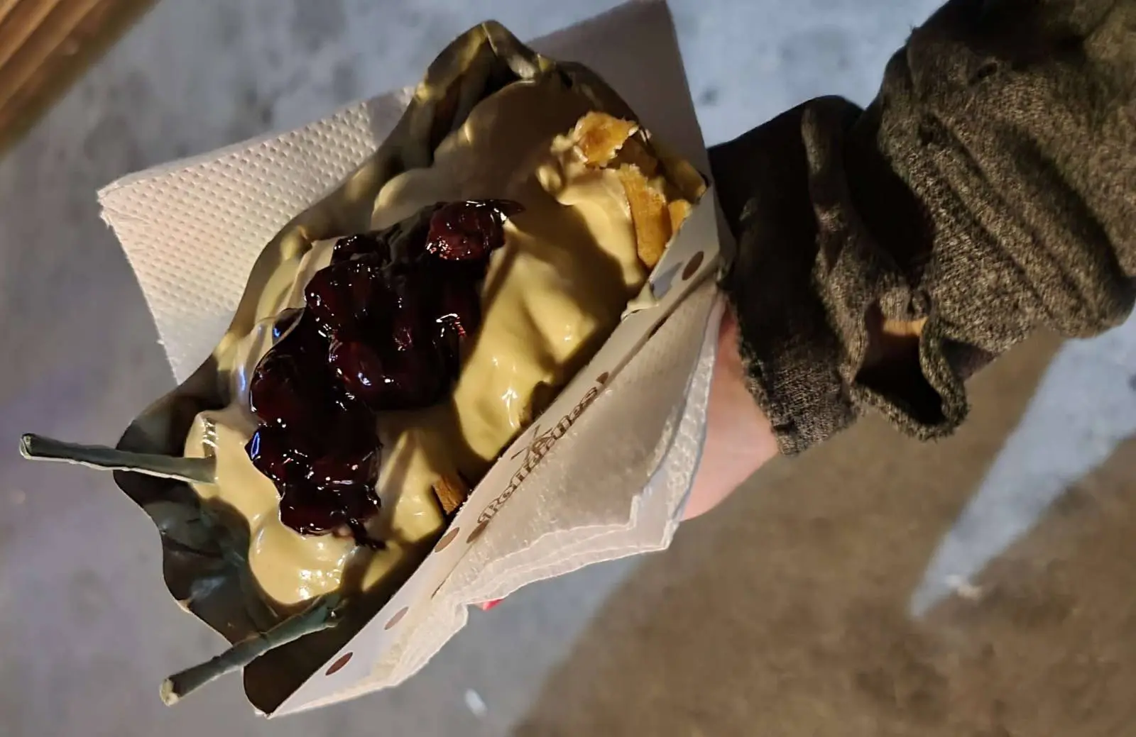 Close-up of a person's hand holding a dessert with waffle pieces, a scoop of ice cream, and a generous topping of dark berry sauce, presented in a paper holder with a napkin. The background is a blurred concrete surface.