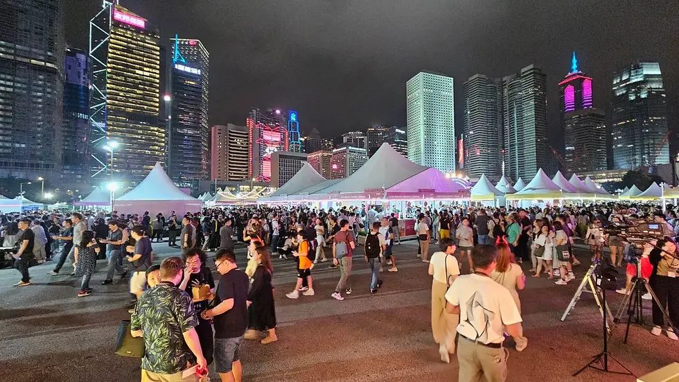 A large crowd gathers at a nighttime outdoor event with white tents and city skyscrapers illuminated in the background.