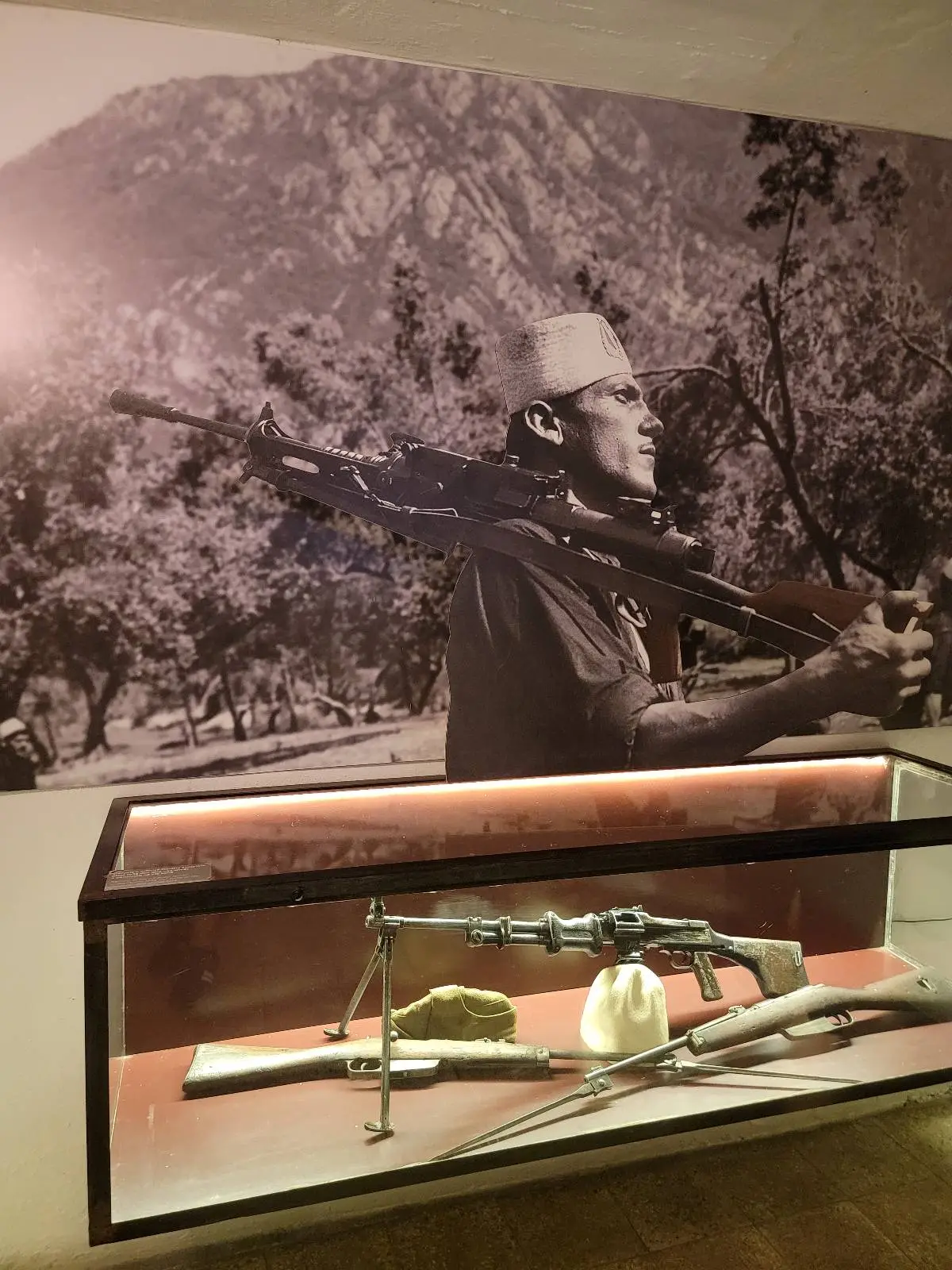A display case contains historic rifles and a helmet. Behind it, a large black and white photograph shows a man carrying a rifle over his shoulder with a mountainous landscape in the background.