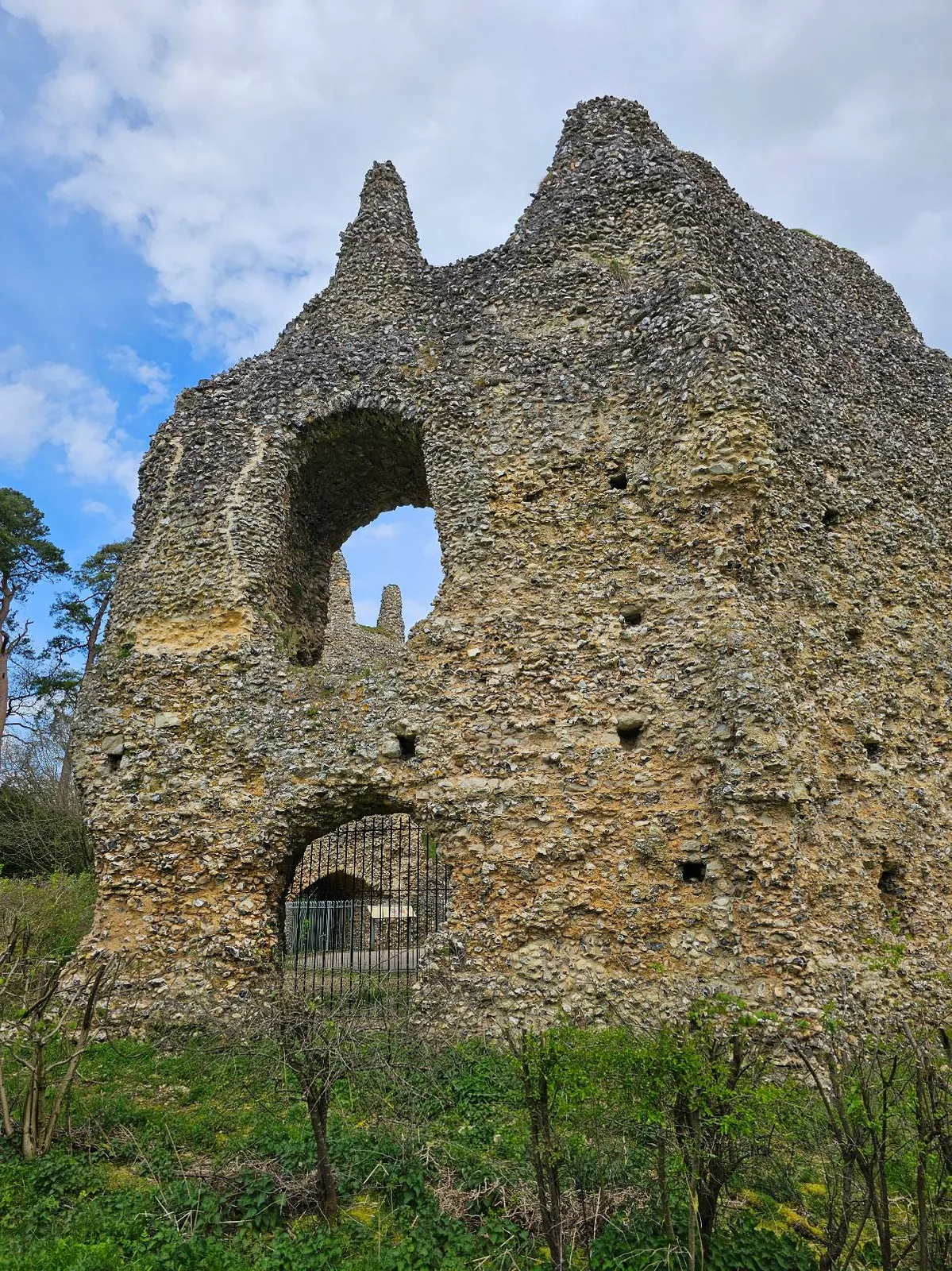 Ruins of an old stone structure with arched openings, set against a backdrop of a blue sky with scattered clouds and greenery in the foreground.