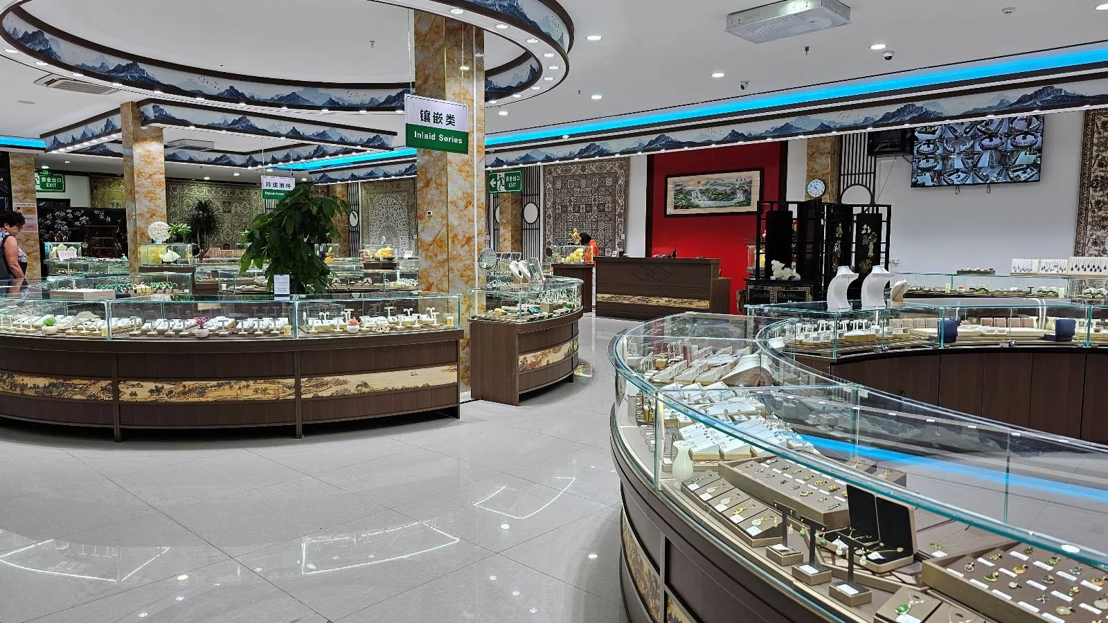 A spacious, well-lit jewelry store with glass display cases filled with various jewelry items, shiny floors, and decorative columns. The store has modern lighting and a luxurious atmosphere.