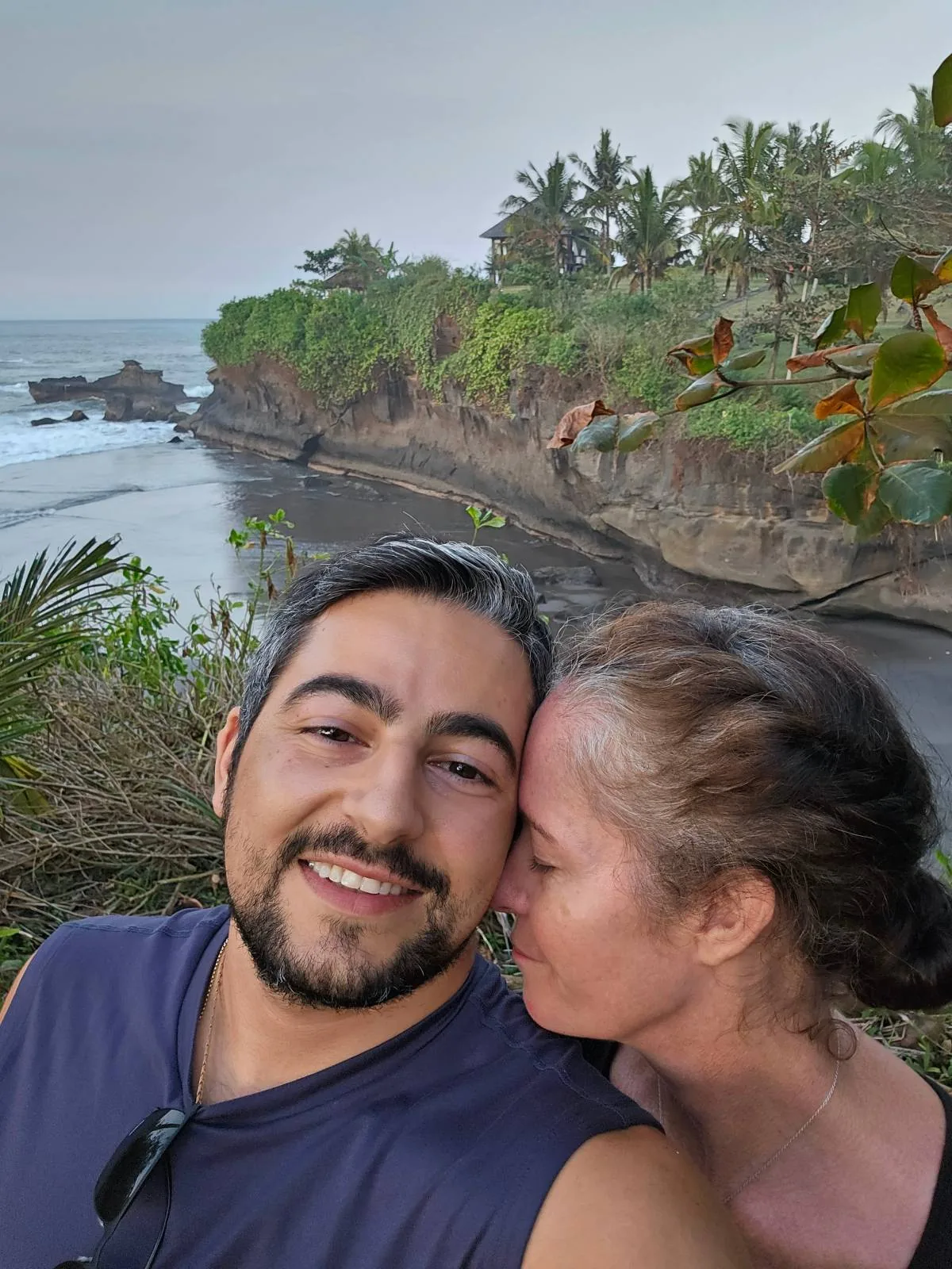 A couple is smiling near a coastal area with lush greenery and ocean waves in the background. The person on the right is leaning affectionately towards the other. The scene is peaceful and natural.