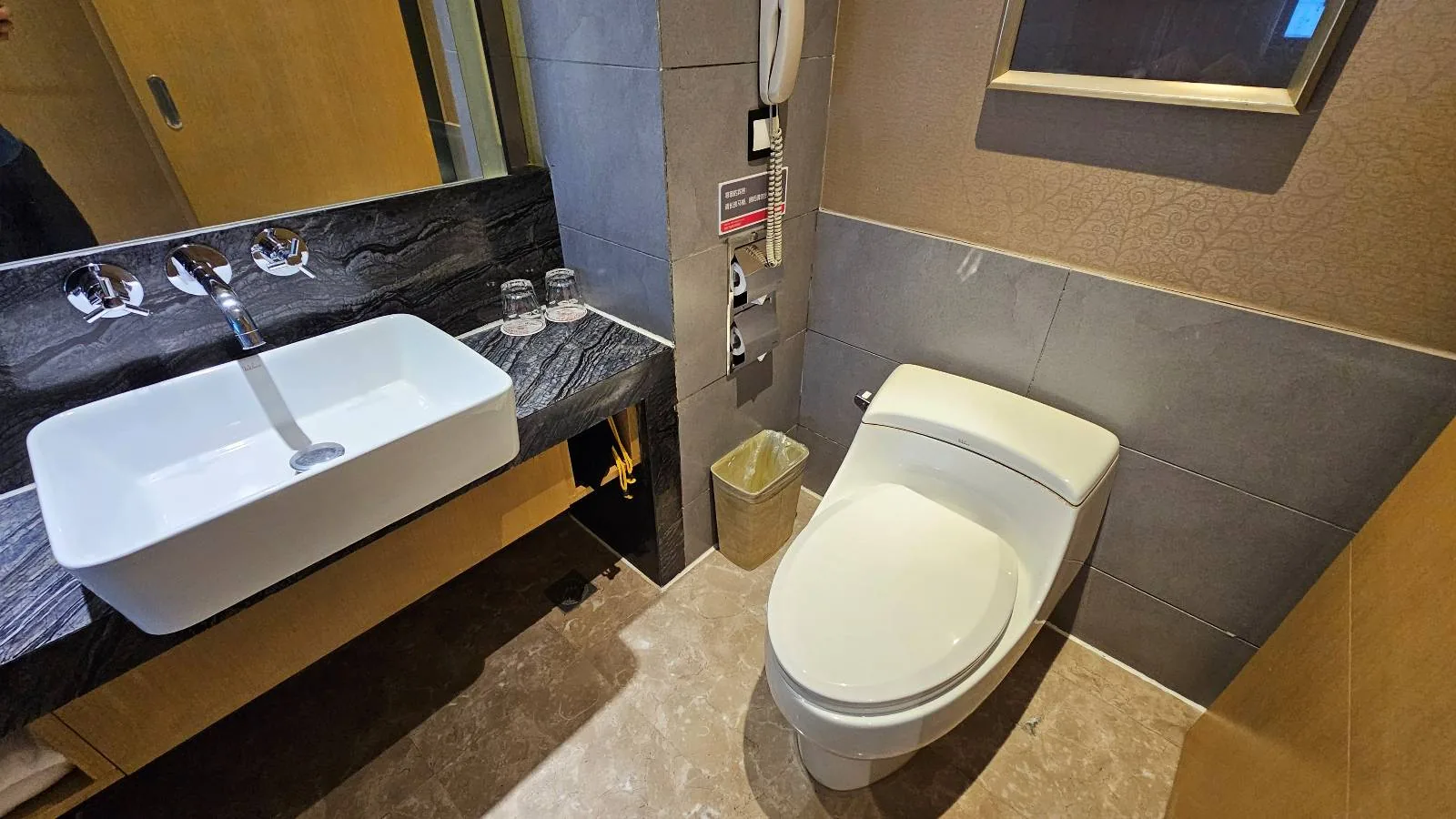 Modern bathroom with a white toilet, a square sink on a black countertop, a mirror, a small trash bin, and a control panel on the wall next to the toilet.
