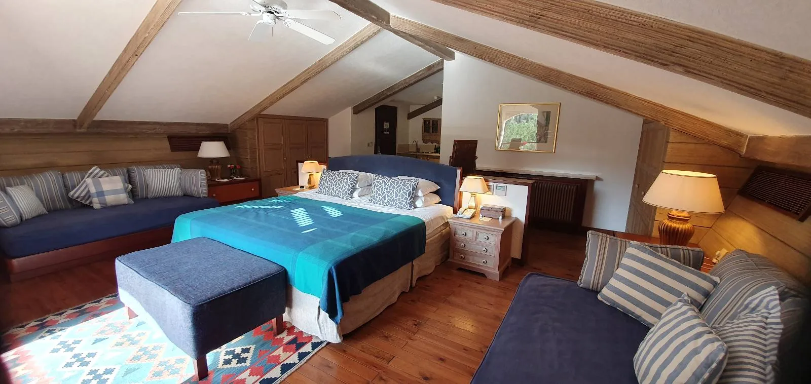 A cozy attic bedroom with a sloped wooden ceiling. It features a large bed with blue bedding, flanked by two nightstands and lamps. There are two cushioned benches, a colorful rug, and art on the walls. Warm lighting creates an inviting atmosphere.