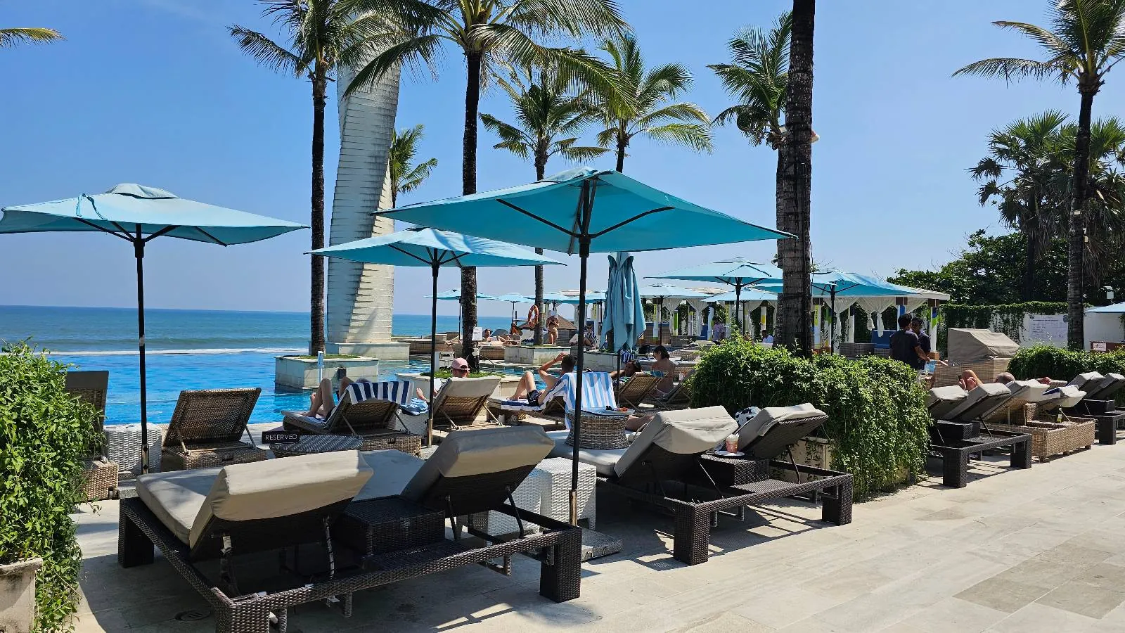 Lounge chairs and blue umbrellas line a poolside area near the beach, with palm trees and a clear blue sky in the background. The ocean is visible just beyond the pool.