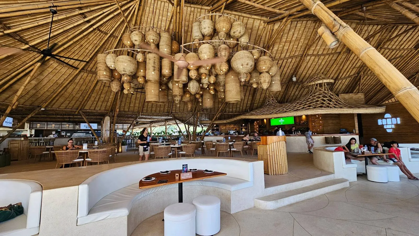 A spacious, open-air restaurant with a thatched roof and large wooden chandeliers. White curved seating and small round tables are scattered around. People are sitting and relaxing. The decor features natural materials and soft beige tones.
