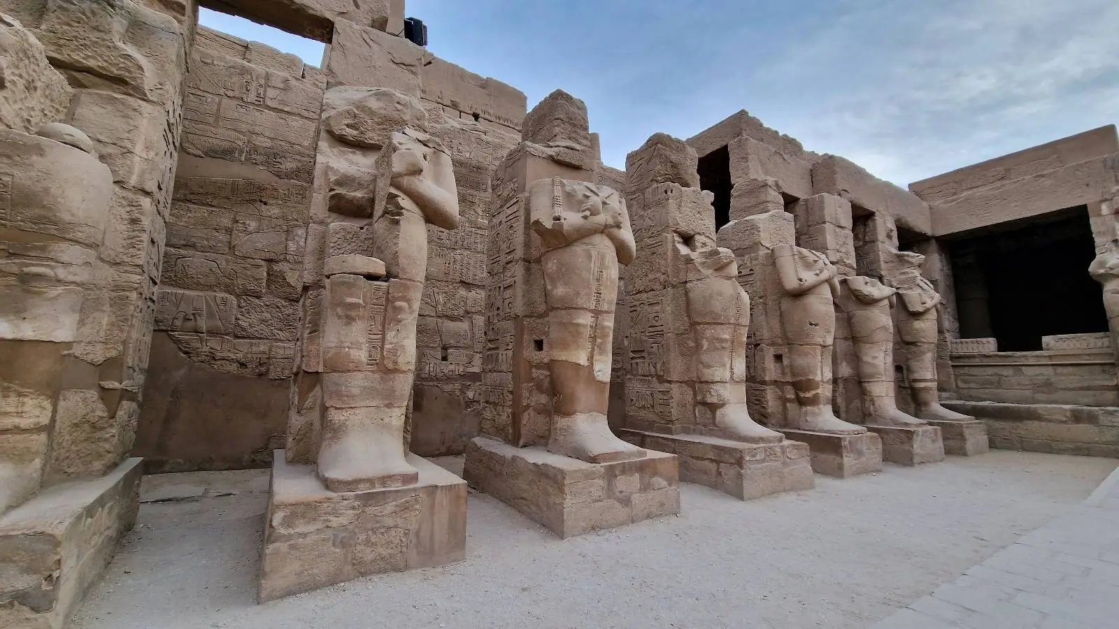 Ancient stone statues lined up in a historical site with a partly cloudy sky above, showcasing detailed carvings and a backdrop of weathered stone walls.