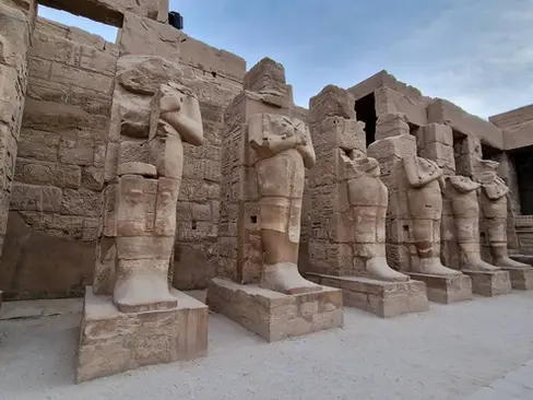 Ancient stone statues lined up in a historical site with a partly cloudy sky above, showcasing detailed carvings and a backdrop of weathered stone walls.