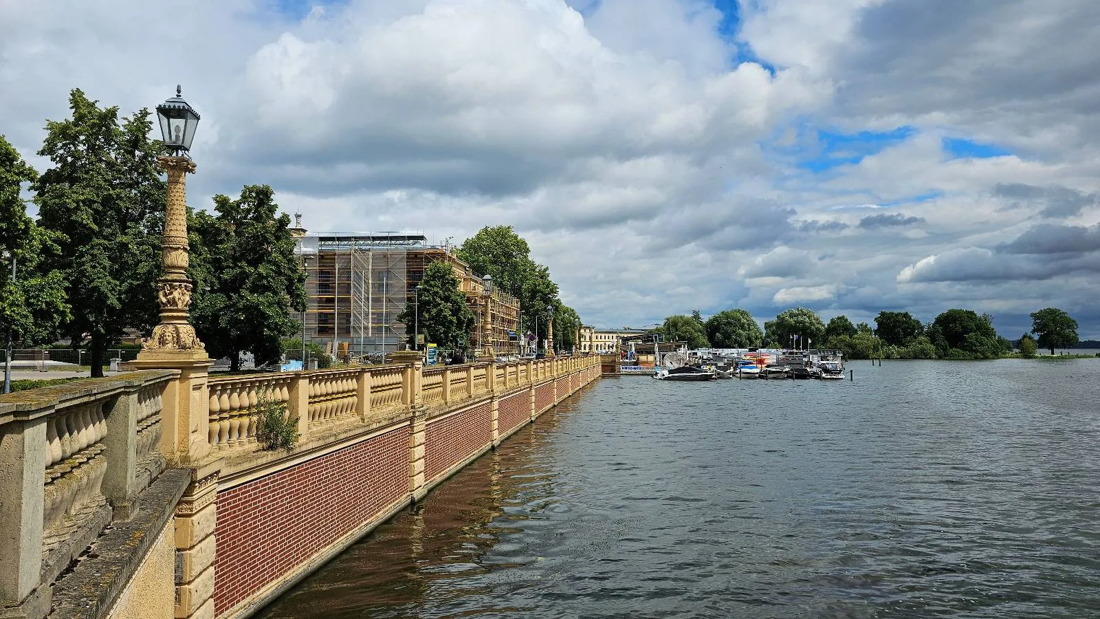 A riverside scene shows a stone and brick walkway with ornate lamp posts, trees lining the path, modern buildings in the background, and boats docked along the water under a partly cloudy sky.