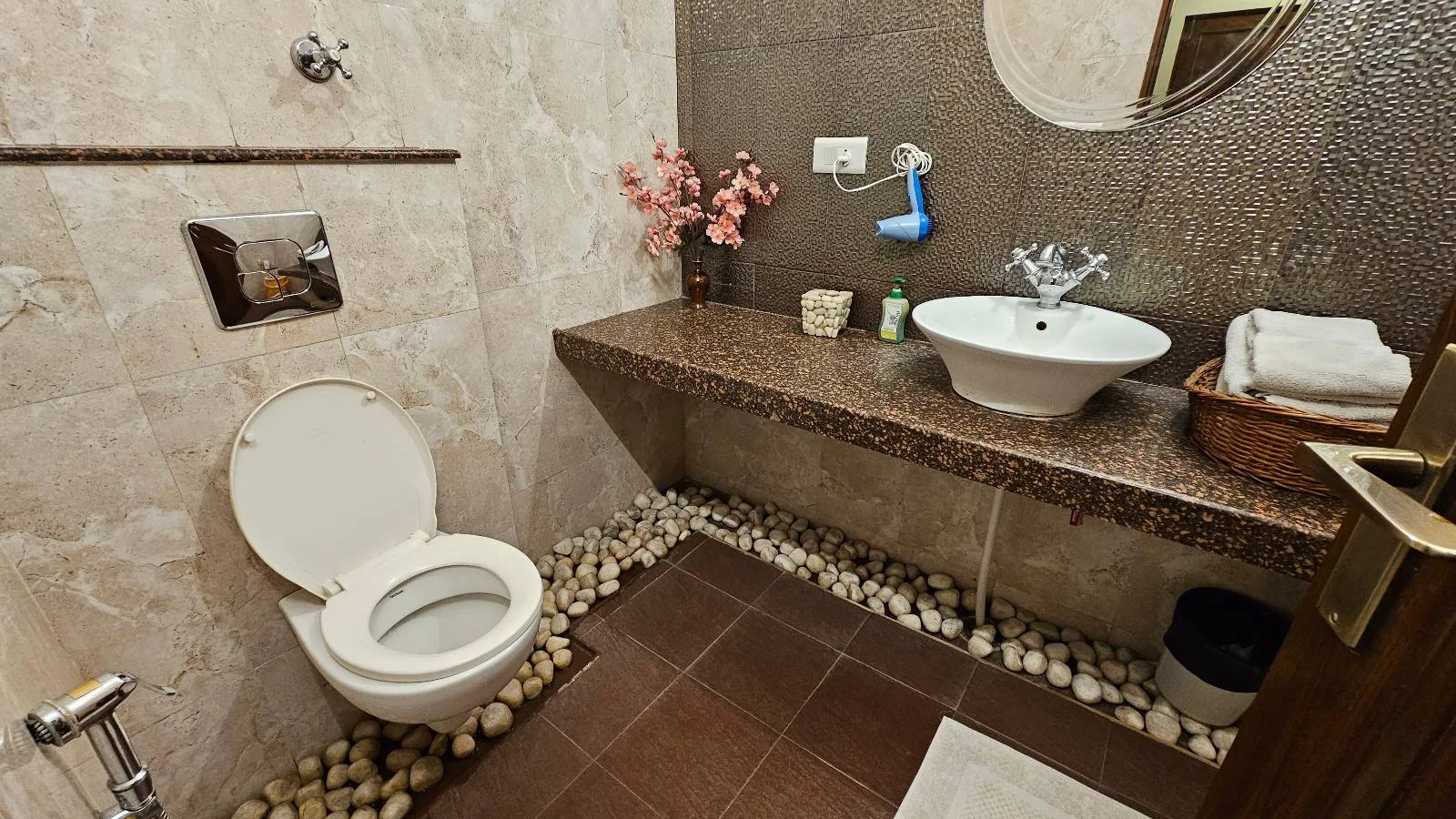 Modern bathroom with a white toilet, open lid, stone accents on the floor, a sink on a granite countertop, pink flowers in a vase, and towels in a wooden basket. Walls are tiled in beige and brown tones.