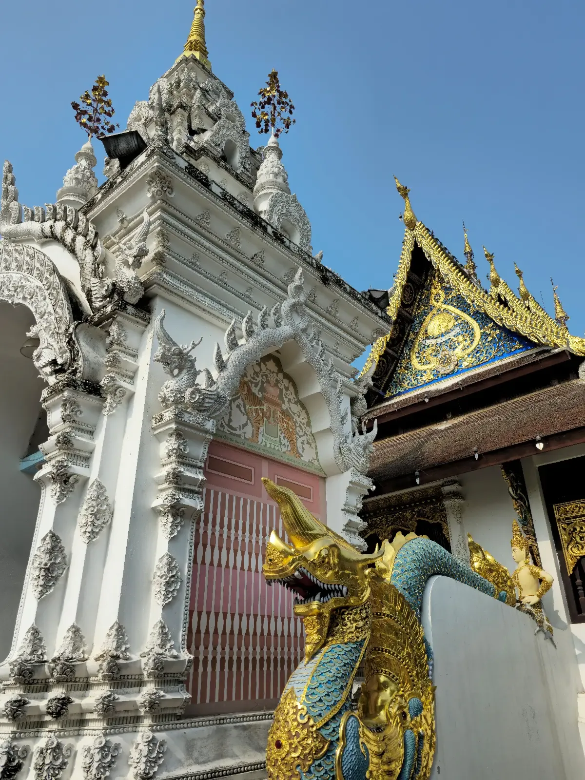 A decorative dragon statue stands in front of an ornate white temple with intricate carvings and a golden roof detail, set against a clear blue sky.