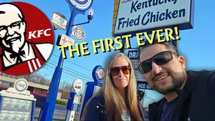 Two people smiling in front of the first KFC sign. Bright blue sky, vintage gas station signs, and "THE FIRST EVER!" text overlay.